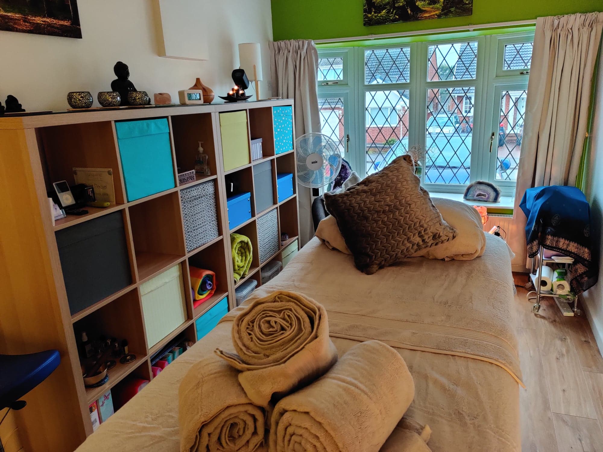 A cozy and well-organized treatment room with a massage table in the center, topped with neatly rolled beige towels and cushions.